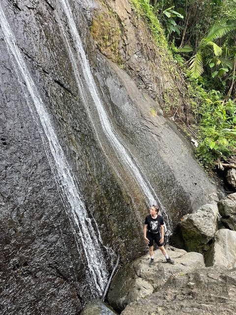 Un caminante al pie de una cascada que baja por una pared de roca oscura cubierta de musgo y helechos